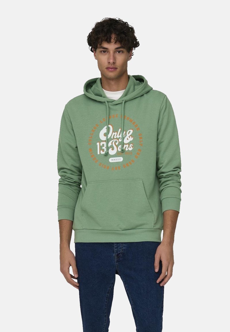 Sudadera verde menta con un gráfico circular de texto en blanco y naranja. Capucha y bolsillo canguro frontal. Combinada con jeans de mezclilla oscura.