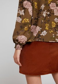 Blouse florale marron avec des fleurs roses et jaunes, dotée de manches bouffantes. Assortie à une jupe en velours côtelé rouille, montrant une texture côtelée.