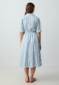 Abito camicia di colore azzurro chiaro, caratterizzato da strisce verticali bianche e blu scuro, maniche corte, cintura in vita e gonna fin sopra il ginocchio.