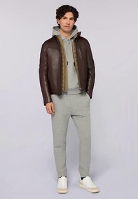 Hnědá kožená bunda s prošitou olivovou podšívkou, šedá mikina s kapucí, šedé tepláky a bílé tenisky. Model stojí s rukama v kapsách.