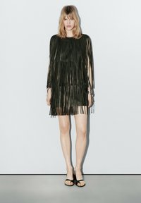 Vrouw met blond haar, gekleed in een zwart minijurkje met franjes en zwarte open teen pumps, staand tegen een effen lichtgrijze achtergrond.