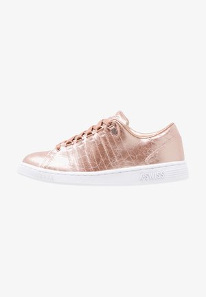 Zapatilla baja de piel texturizada en tono rosa metálico, con cordones planos y suela de goma blanca con la marca "K-Swiss" en el talón.