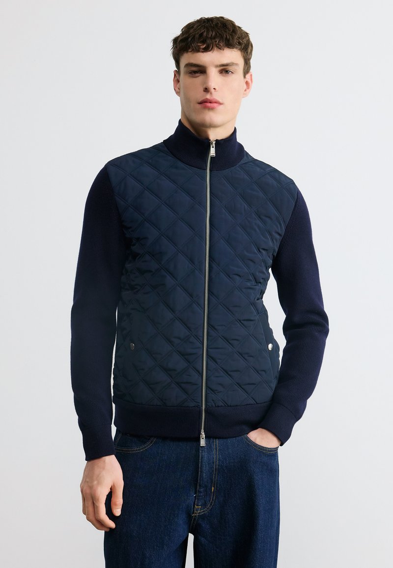 Νέος άνδρας που φοράει μπλε σκούρο μπουφάν με φερμουάρ, πλεκτά μανίκια και γιακά, συνδυασμένο με σκούρο μπλε τζιν, στέκεται μπροστά σε απλό φόντο.