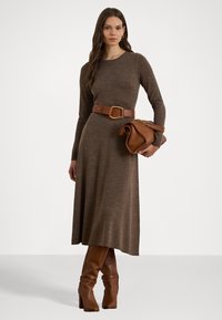 Bruine gebreide midi-jurk met lange mouwen en een ronde halslijn, cinched in de taille met een brede bruine riem en geaccessoriseerd met een handtas.