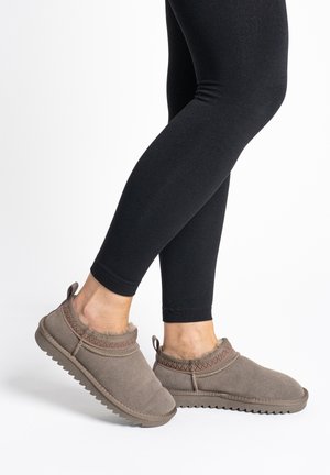 Persona che indossa leggings neri e pantofole taupe con fodera in pile e suole in gomma testurizzata, in piedi su una superficie bianca.