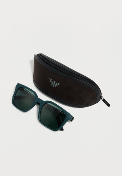 Σκούρα τιρκουάζ γυαλιά ηλίου τετραγωνικού σχήματος με πράσινους φακούς τοποθετημένα δίπλα σε μαύρη θήκη με φερμουάρ και ασημένιο λογότυπο, πάνω σε λευκό φόντο.