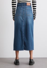Gonna midi in denim blu scuro con due tasche posteriori, uno spacco frontale e una patch con il logo. Tessuto morbido e silhouette dritta. Scarpe nere visibili.