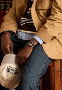 Montre en cuir marron avec un cadran noir, accents dorés, et un blazer beige avec un pull à rayures. Chemise bleu clair et jeans en denim. Casquette blanche en dessous.