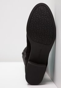 Chaussure en cuir suédé noir avec une semelle en caoutchouc texturé, arborant le logo Calvin Klein sur le talon et une surface lisse. Le design est bas.