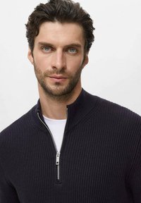 Donker marineblauwe geribde zip-up sweater met hoge kraag en zilveren ritsdetail. Gdragen over een witte ronde hals t-shirt. Glad van textuur.