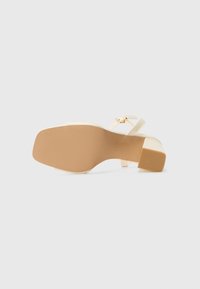 Beige hælhæl med en cremefarvet rem og spænde detalje. Den teksturerede sål er lys brun med et blokhælsdesign.