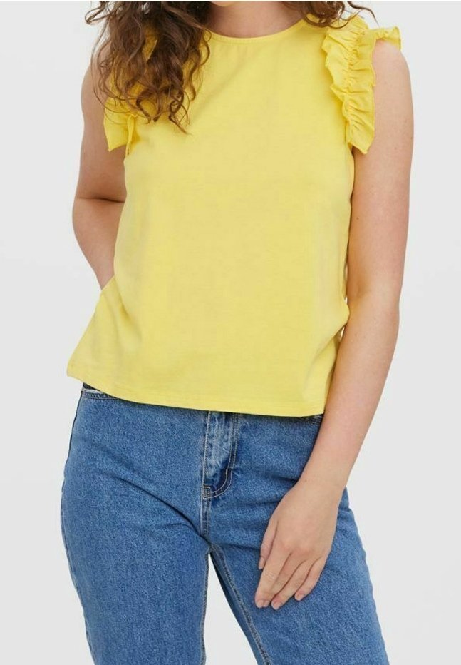 Femme portant un haut jaune sans manches avec des épaules à volants et un jean bleu, debout, une main dans la poche et l'autre sur le côté.