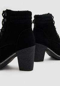 Botines de ante negros con cuellos acanalados acolchados, tacones en bloque y cierres de cremallera lateral sobre un fondo blanco liso.