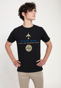 Černé bavlněné tričko s grafickým designem, na kterém je modrý a zlatý text "PAN AM" a silueta letadla, spolu s logem.