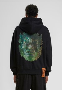 Sweat à capuche noir avec un grand graphique coloré d'un visage au dos, avec une surface texturée et une étiquette orange en bas.