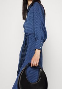 Blå mönstrad klänning med bälte, knappade manschetter och en svart halvmånehandväska med texturerad yta. Slätt tyg, strukturerad silhuett.