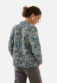 Chaqueta acolchada en azul con un patrón floral en crema y amarillo. Cuenta con cuello, mangas largas y un ribete en contraste. La textura parece suave.