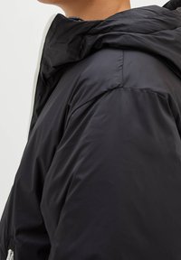 Veste matelassée noire à capuche, avec une texture matelassée et une silhouette élégante. Le tissu semble lisse avec des détails matériels minimaux.