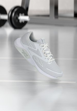 Lys grå og hvid sportssko med meshstof og logo, placeret i et fitnessmiljø med slørede vægte i baggrunden.