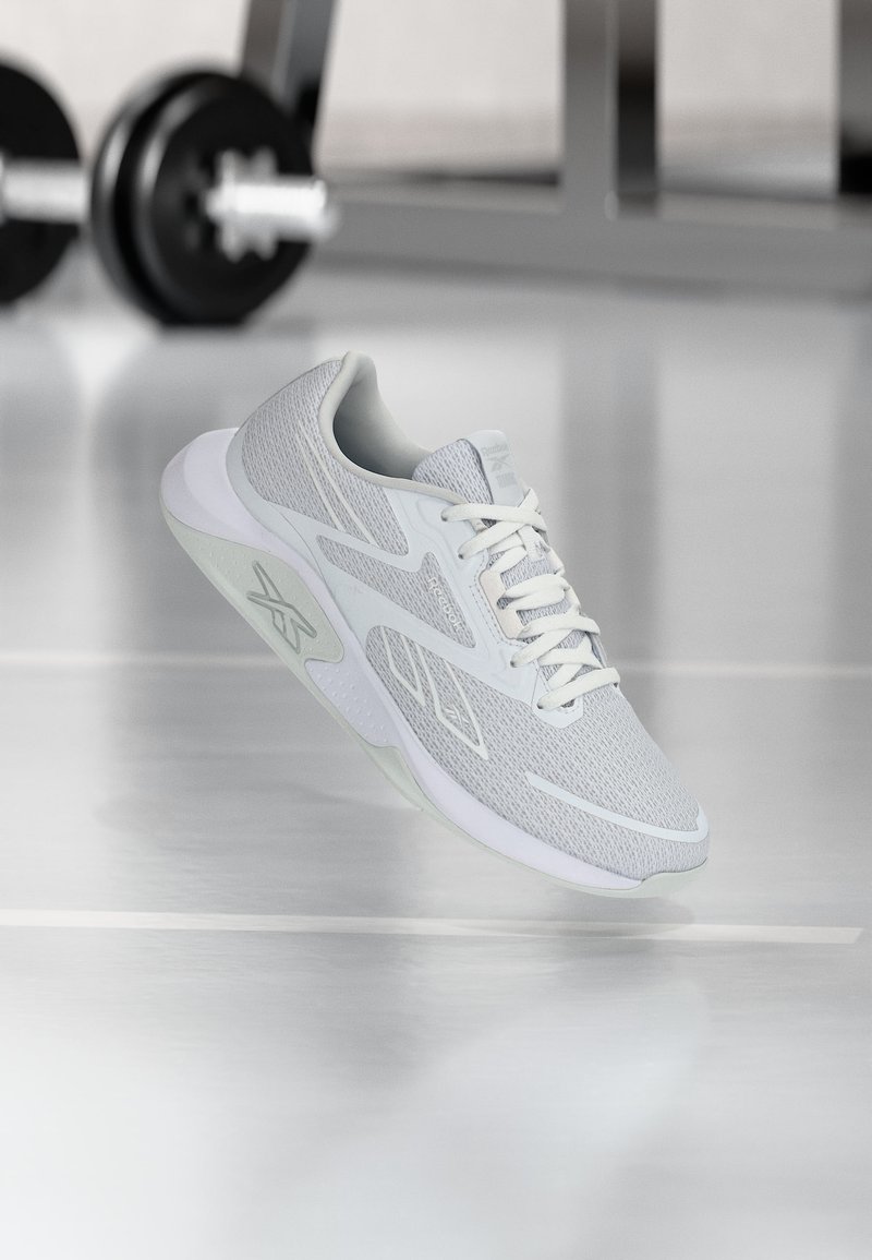 Αθλητικό παπούτσι ανοιχτό γκρι και λευκό με υφασμάτινο πλέγμα και λογότυπο, τοποθετημένο σε γυμναστήριο με θολό φόντο βαρών.