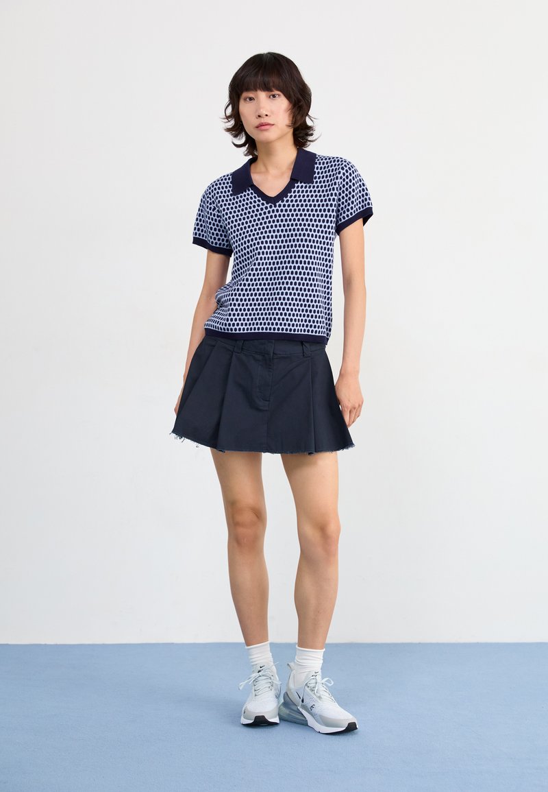 Camisa de polo azul marino con lunares blancos, de manga corta y escote en V, combinada con una falda acampanada azul marino oscura y zapatillas claras.