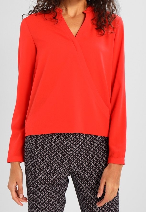 Blusa rossa a maniche lunghe con scollo a V e design avvolgente. Realizzata in tessuto morbido; abbinata a pantaloni neri a fantasia con piccoli disegni bianchi.
