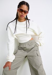 Bela skrajšana mikica z ribastim notranjim slojem in elastičnimi manšetami, v kombinaciji z ohlapnimi bež cargo hlačami, ki imajo logo in žep. Smooth as in white cropped sweatshirt with ribbed inner layer and elastic cuffs, paired with loose-fitting beige cargo pants featuring logo and pockets. 