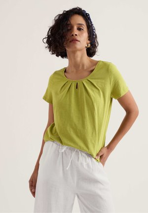 Vrouw met krullend haar die een limegroen T-shirt met korte mouwen en witte broek met trekkoord draagt, poserend tegen een effen achtergrond.