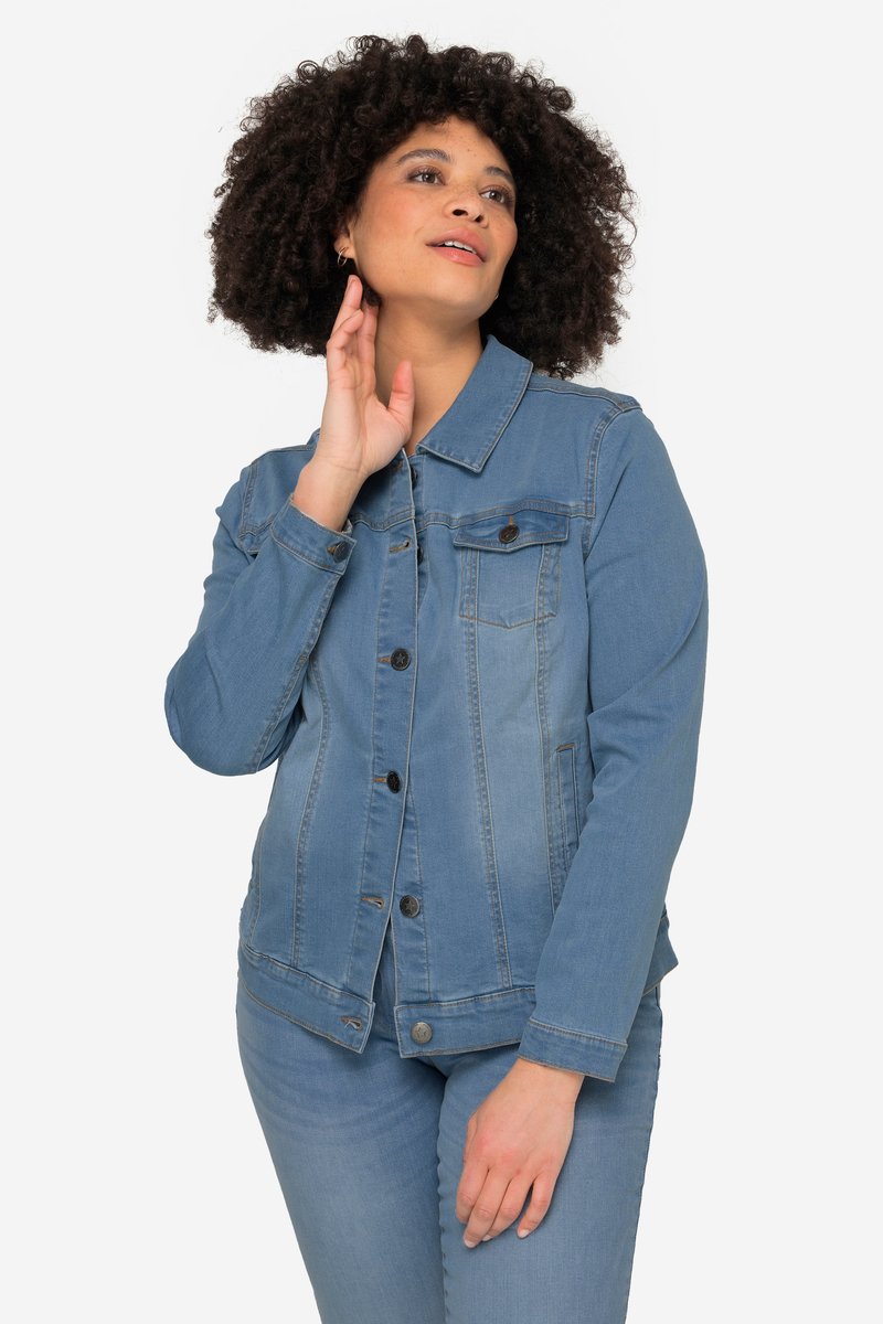 Kvinde med krøllet hår iført en lyseblå denimjakke med knapper og jeans, der rører ved sin nakke og kigger opad til siden.