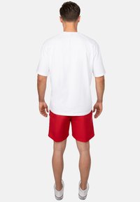 T-shirt bianco in cotone a maniche corte, abbinato a pantaloni rossi. Presenta una vestibilità comoda e un design semplice, con cuciture visibili sui bordi.