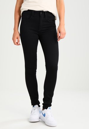 Persoon draagt nauwsluitende zwarte spijkerbroek en witte sneakers met blauw Nike-logo, staand tegen een effen lichte achtergrond.