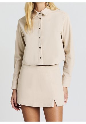 Chemise beige côtelée à manches longues boutonnée et mini-jupe taille haute assortie fendue sur le devant, portée par une personne sur un fond uni.
