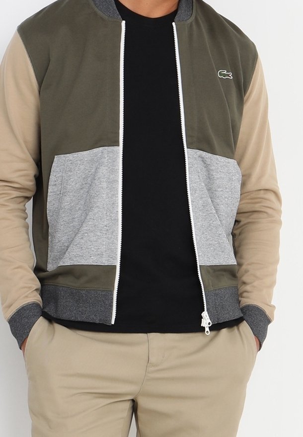 Homme portant une veste color-block avec des parties olive, beige et grises, sur une chemise noire, les mains dans les poches d'un pantalon beige.