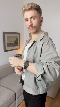 Μαύρο ρολόι χειρός σε ένα χέρι, με στρογγυλή πρόσοψη και καθαρές ενδείξεις. Το άτομο φοράει ένα ανοιχτό γκρι σακάκι πάνω από ένα λευκό πουκάμισο.