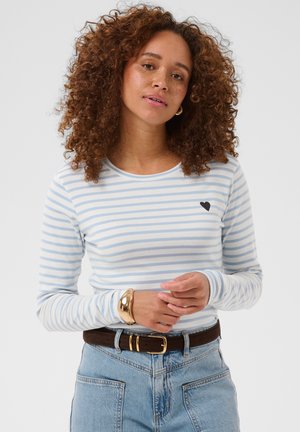 Kvinde med krøllet hår iført en lyseblå og hvidstribet langærmet skjorte, denim jeans, brun bælte og gyldt armbånd mod en ensfarvet baggrund.