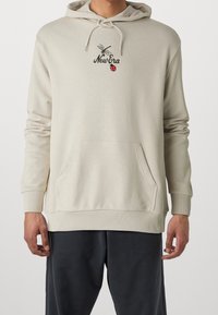 Felpa beige con tasca frontale e cordini, caratterizzata da testo "New Era" con una grafica di una libellula e una coccinella sul petto, indossata con pantaloni scuri.