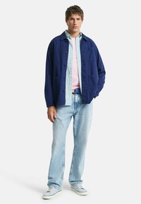 Chaqueta navy de botón sobre una camisa de rayas azul claro y una camiseta rosa, combinada con jeans de denim claros y zapatillas blancas.