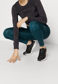 Mustapaitainen pitkähihainen toppi, jossa on Puma-logo, teal-väriset leggingsit ja mustat urheilukengät valkoisilla yksityiskohdilla. Malli kyykistyy ja koskettaa maata.