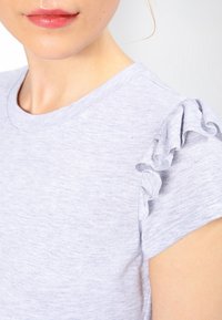 Světle šedé tričko s krátkými rukávy a volánkovými detaily na ramenou, vyrobené z měkké látky s jemnou texturou.