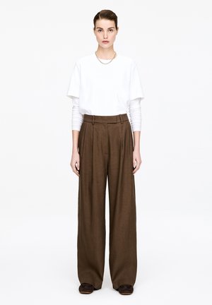 Kvinde iført lagdelte hvide toppe med lange og korte ærmer, brune vidbenede bukser med høj talje og mørke sko, stående mod en hvid baggrund.