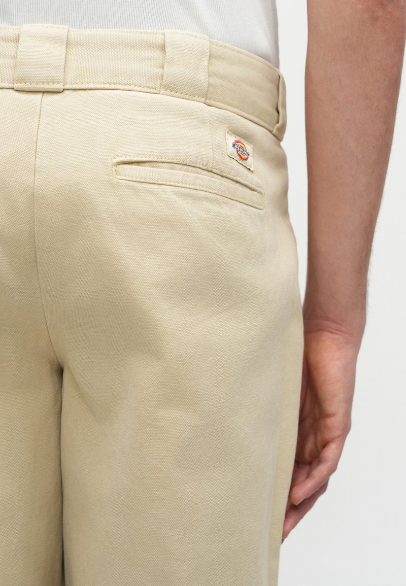 Pantaloni beige con una tasca a filetto sul retro e un piccolo logo vicino alla tasca, indossati da una persona con il braccio rilassato al fianco.