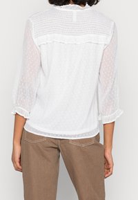 White blouse with semi-sheer, textured fabric featuring small dots. Ruffled accents on sleeves and neckline, relaxed fit, and plain back.