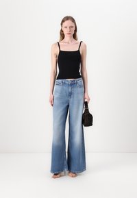 Svart ribbet tank topp og lyseblå vide jeans med gradvis falming. Holder en liten brun håndveske, satt mot en ensfarget bakgrunn.