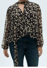 Blouse noire transparente avec imprimé marguerites, manches longues, fermeture à nouer au col et ourlet arrondi. Design à boutons.