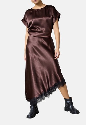 Kvinde iført en skinnende brun satin top og asymmetrisk nederdel med sort blonderkant, kombineret med sorte læderankelstøvler.