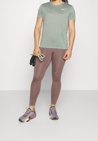 Šedé tričko s krátkým rukávem Nike, těsné hnědé legíny a fialové sportovní boty. Osoba drží cvičební gumy. Hladký materiál, aerodynamický design.