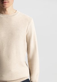 Lys beige genser med teksturert strikkemønster, ribbestrikkede cuffs og hem, samt liten logo detalj på brystet. Vanlig passform og myk stoff.