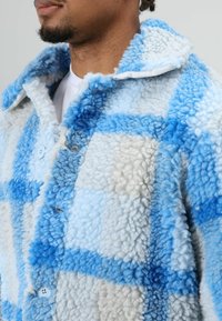 Fleece jacket in light blue, dark blue, and cream plaid pattern. Features soft texture, button closure, and wide collar design.