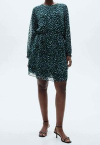 Vestido verde con estampado, de manga larga, con cintura elástica y una silueta fluida. Combinado con sandalias negras de tiras.