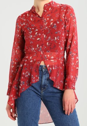 Blouse rouge à manches longues à motif floral avec boutons sur le devant et ourlet asymétrique, associée à un jean bleu taille haute.